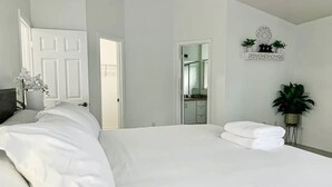 A bedroom with a large bed, a bedside table with a lamp, a door leading to a bathroom, and a wall-mounted shelf with decorative items.