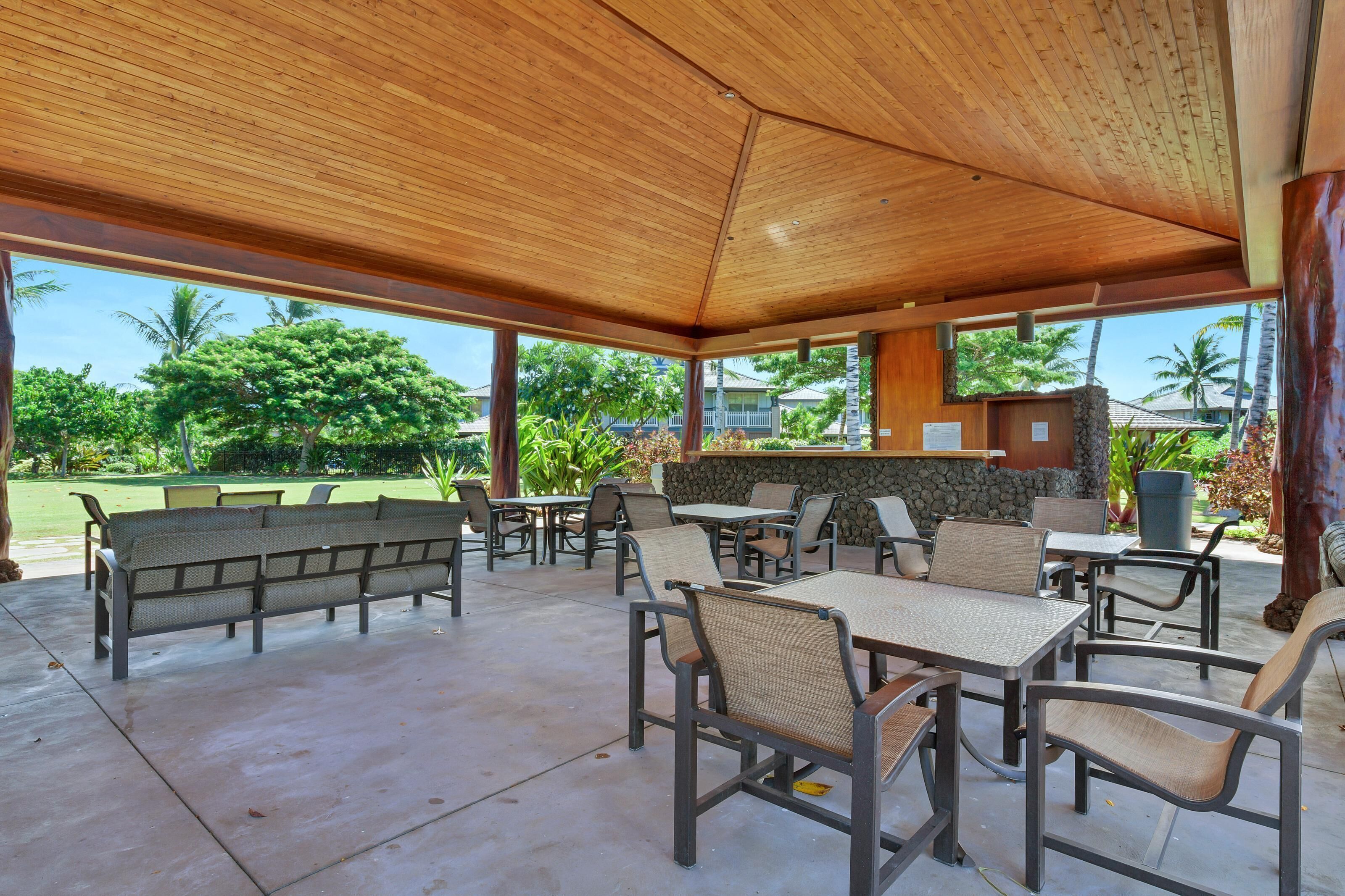 An outdoor covered patio with seating and tables, a stone wall, and a view of greenery and buildings.