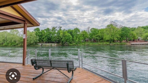 Una terraza de madera con un banco de metal que da a un río, rodeada de árboles y un cielo nublado.