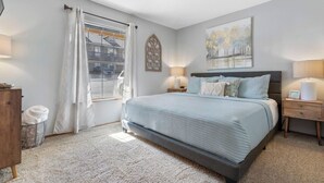 Un dormitorio con cama, ventana con cortinas, mesita de noche con lámpara y una obra de arte colgada en la pared.