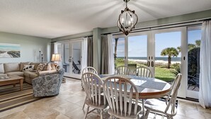 A dining area with a table and chairs, a sofa, and a view of the beach through large sliding doors.