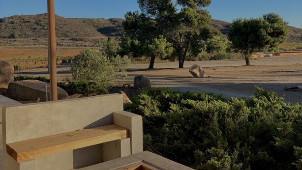 Un espace extérieur aménagé avec des sièges, doté d’un foyer en béton, entouré d’arbustes et d’arbres, et surplombé d’une pergola en bois.