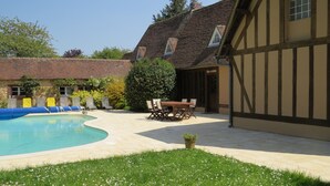 A swimming pool with a curved edge, surrounded by a grassy area with white flowers. There is a wooden table and chairs in the background, and a building with a tiled roof and wooden beams. - Normandy vacation home with swimming pool 12km from Verneuil sur avre (Mesnils sur Iton)