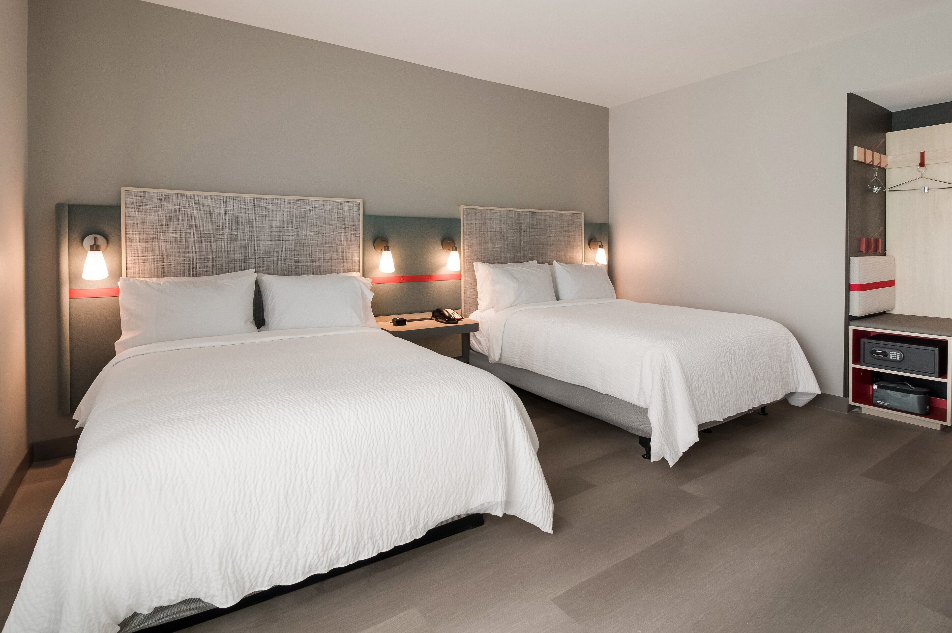 A hotel room with two beds, each with a headboard and bedside tables. There is a wardrobe with a mirror and a shelf with items on it.