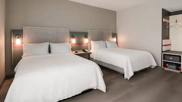 A hotel room with two beds, each with a headboard and bedside tables. There is a wardrobe with a mirror and a shelf with items on it.