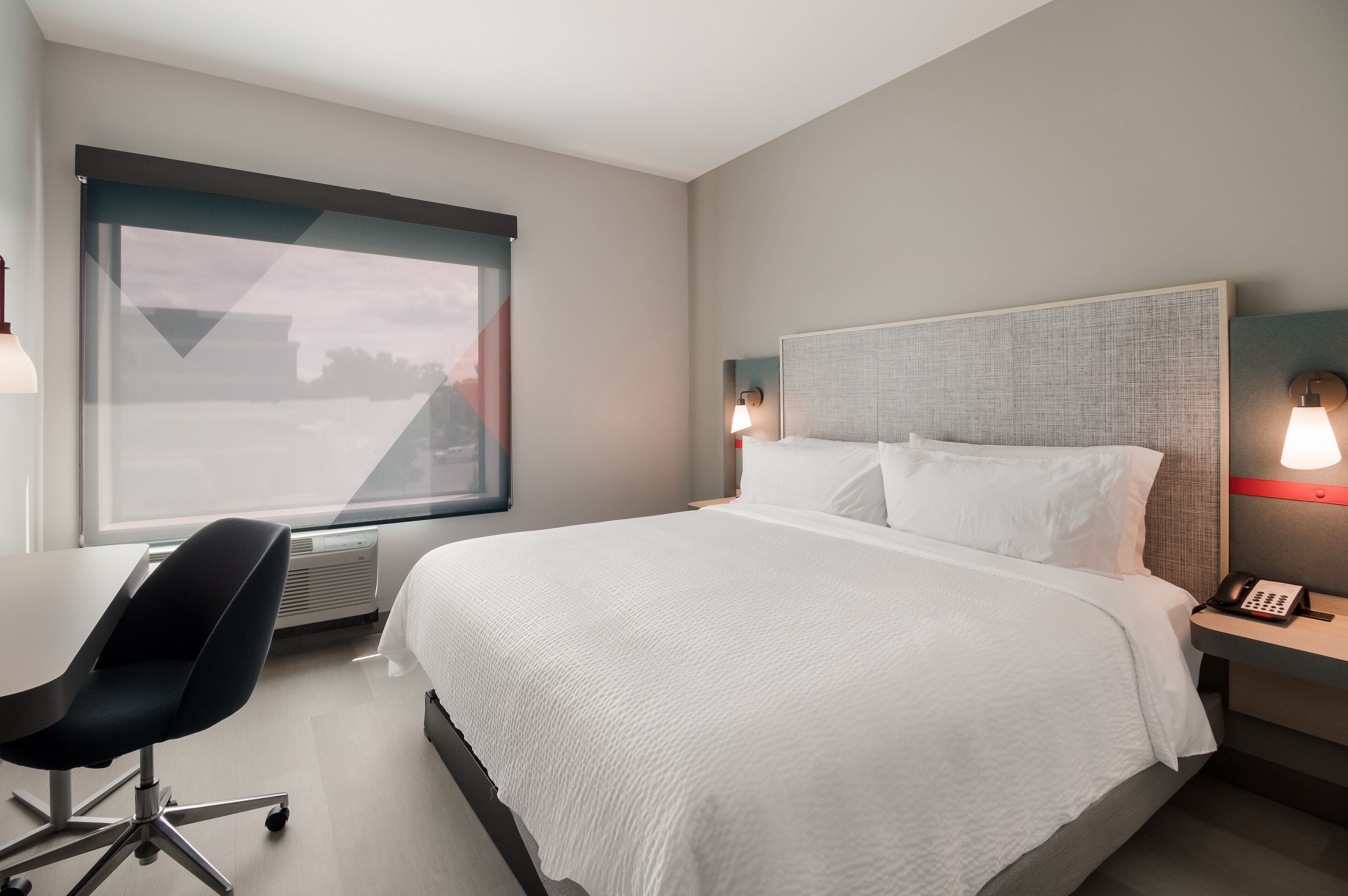 Habitación de hotel con una cama grande, un escritorio y una silla. Se ve un ventanal con vistas al exterior.
