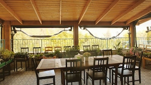 Een terras buiten met houten tafels en stoelen, omgeven door potplanten en een metalen hekwerk.