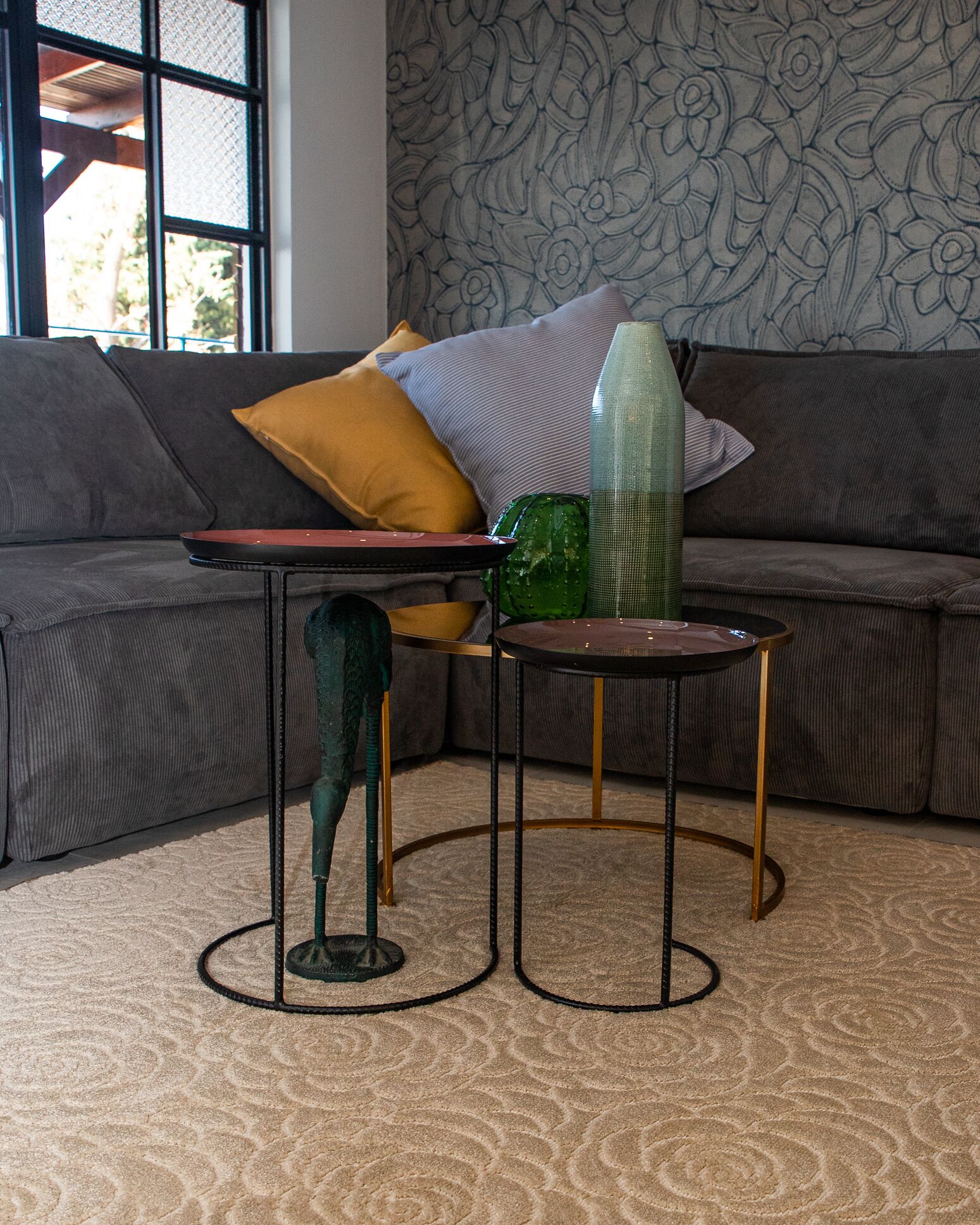 Een hoek van een kamer met een grijze bank, een behang met patroon en twee kleine tafels met metalen poten.