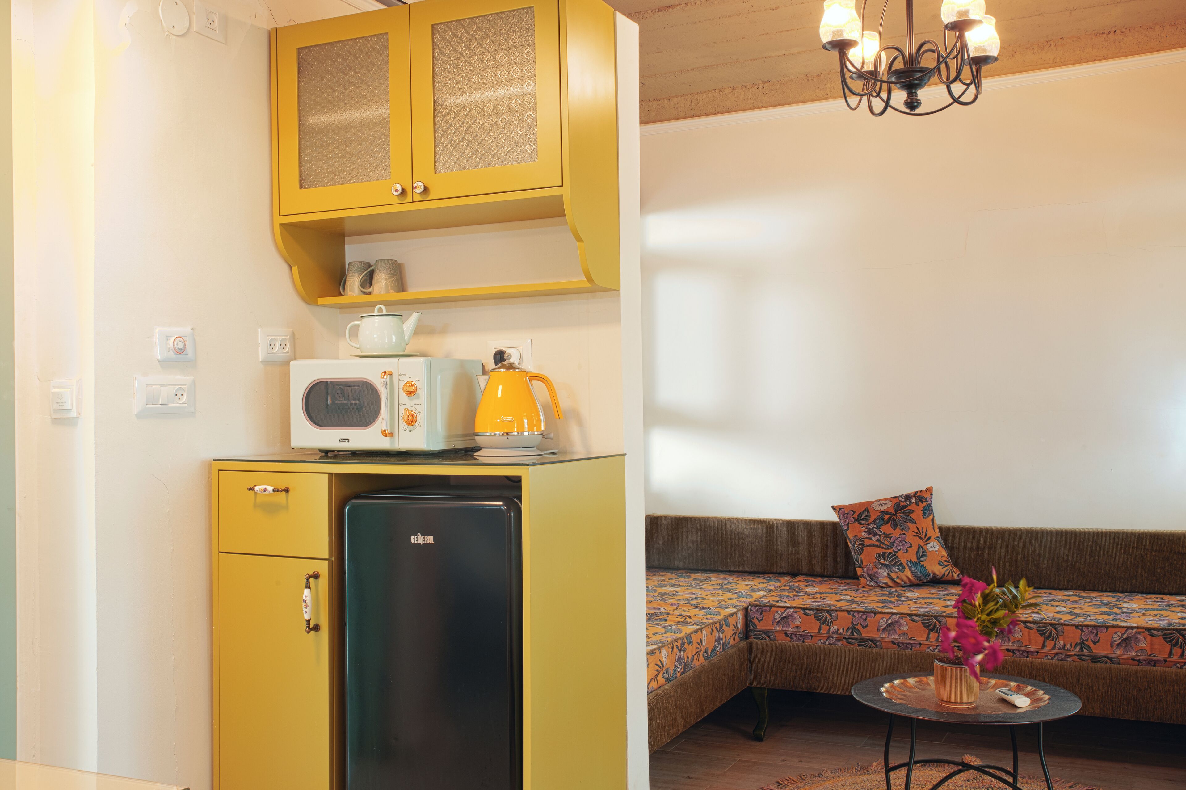 A compact kitchen with a refrigerator, microwave, and kettle, and a cozy seating area with a patterned sofa and a small table with a vase.