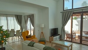 Un ampio soggiorno con divano, poltrone, tavolino e televisore. È presente anche una zona pranzo con sedie e tavolo, e un balcone con mobili da esterno.