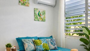 Un dormitorio con colcha color turquesa, almohadas con motivos tropicales, una mesita redonda con una planta y obras de arte en la pared.