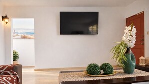 Una sala de estar con un televisor montado en la pared, una mesa de centro con objetos decorativos, un sofá y vistas a la playa a través de una puerta abierta.