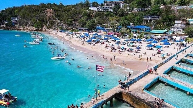 Una playa con agua turquesa cristalina, un muelle con gente caminando y sombrillas de playa.