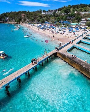 A beach with clear turquoise water, a pier with people walking, and beach umbrellas. - Crash Boat Beach Oasis Spacious 4-Bed/3-Bath Beach House Located in the center. (Aguadilla)