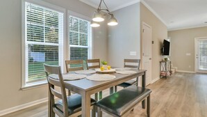 Un coin repas avec une table en bois, quatre chaises et un banc. Il y a deux fenêtres équipées de stores, un luminaire au plafond et un téléviseur fixé au mur.