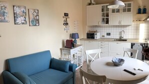A cozy living room with a blue sofa, a white dining table, and a kitchen area with white cabinets and a microwave. - In the heart of Tuscany, beautiful garden with swimming pool, Casa di Ele awaits you (Gambassi Terme Firenze)