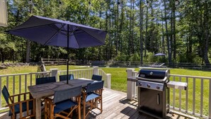Um deck externo com mesa, cadeiras, churrasqueira e um guarda-sol grande.
