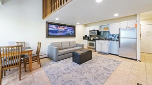 A modern living room with a grey sofa, wooden dining table, and chairs. A black ottoman is in the center. The kitchen area includes a microwave, oven, and refrigerator.