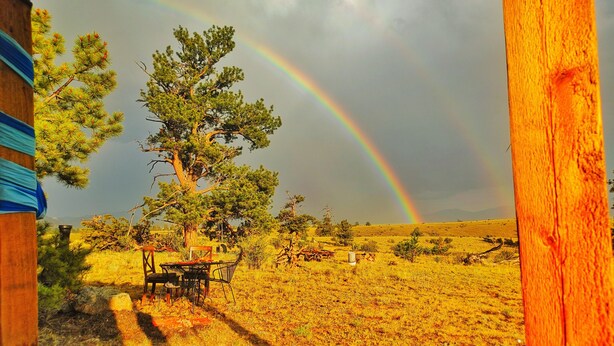 A wooden deck with a table and chairs, a tree, and a rainbow in a grassy field.