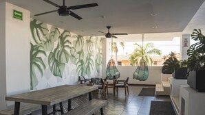 Ein Restaurant mit modernem Design, das ein großes Wandbild mit tropischen Blättern, verschiedene Sitzbereiche mit Tischen und Stühlen, Deckenventilatoren und Hängestühle zeigt.