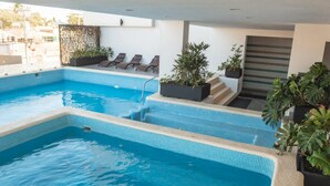 Ein moderner, überdachter Swimmingpool mit klarem, blauem Wasser, einem kleinen Wasserfall und einer Sitzecke mit Liegestühlen und Pflanzen in Töpfen.