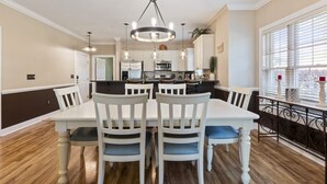 Un coin repas avec une table et des chaises blanches, un lustre et, au fond, une vue sur la cuisine.