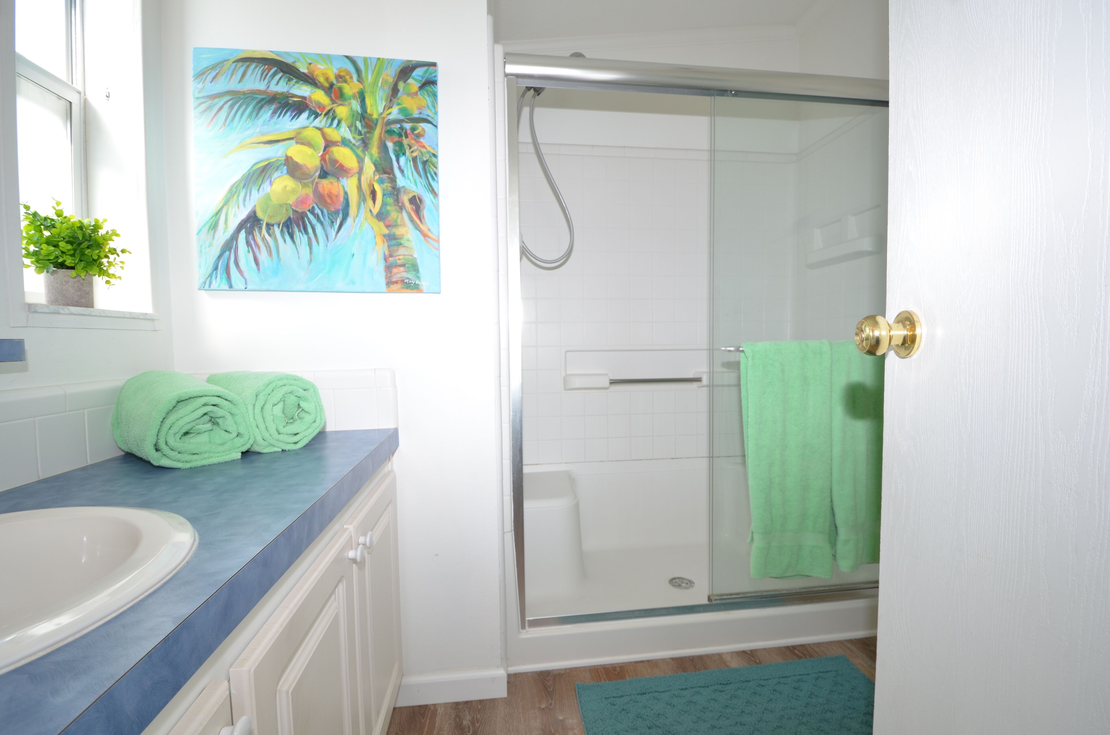 A bathroom with a glass shower enclosure, a white sink, and a painting of palm trees.