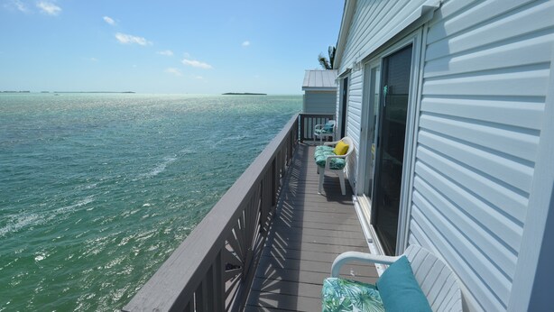 A balcony with a view of the ocean, a small deck area with chairs and cushions, and a building with a sliding glass door.