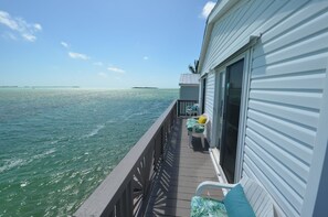 A balcony with a view of the ocean, a small deck area with chairs and cushions, and a building with a sliding glass door. - House of the Rising Sun - VO479 (Cudjoe Key)
