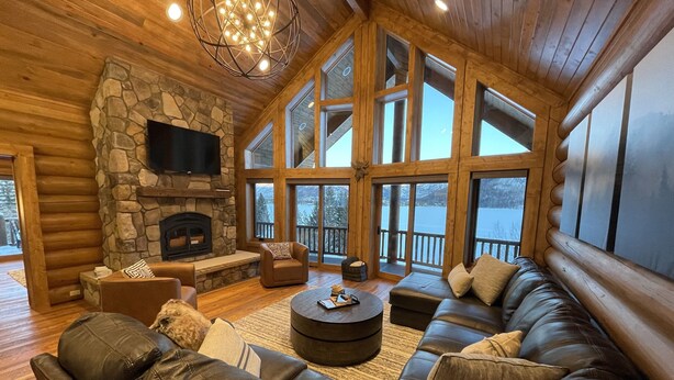 A log cabin interior with a stone fireplace, leather sofas, a flat-screen TV, and large windows offering a view of a snowy landscape. - Best Mtn/Lake views!!! Lodge on esteemed Lakeside Drive! Hot tub! Fido friendly! (Grand Lake)