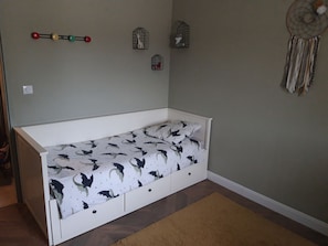 A single bed with a patterned bedspread, a brown rug, and wall decorations including birdcages and a dreamcatcher.