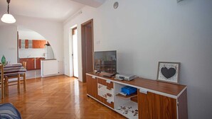 Een moderne woonkamer met een houten vloer, een tv op een houten dressoir, een eethoek met een geruit tafelkleed, en een keuken op de achtergrond.
