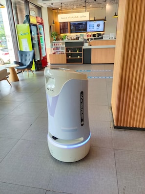 A self-service coffee machine with a digital display, a seating area with chairs, and a menu board in a cafe setting.