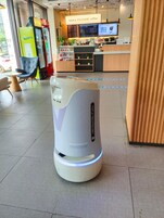A self-service coffee machine with a digital display, a seating area with chairs, and a menu board in a cafe setting.