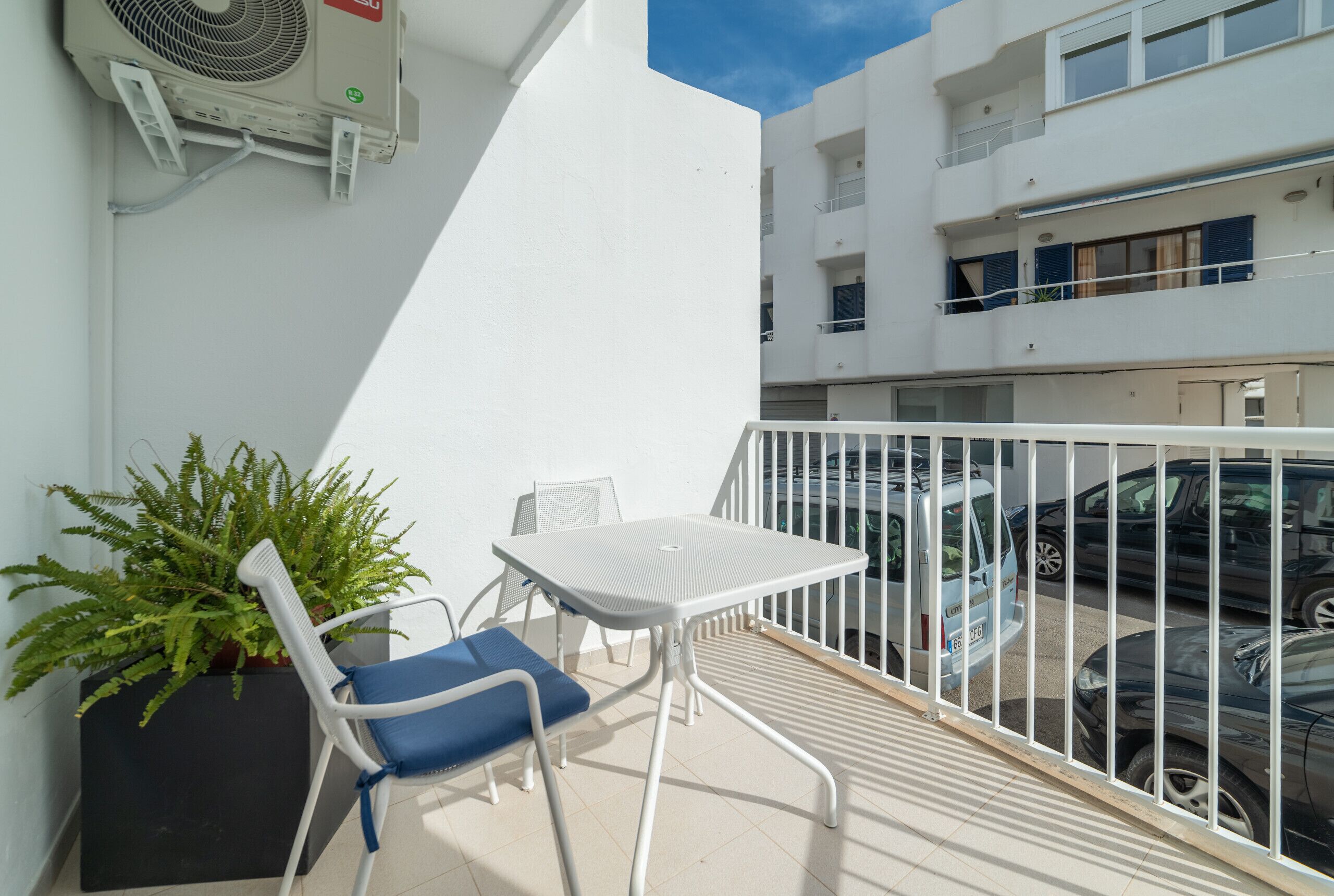 Un balcón con una mesa y una silla blancos, una planta en maceta y vistas a coches aparcados.