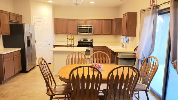 A modern kitchen with wooden cabinets, a stainless steel refrigerator, a microwave, an oven, and a sink. There is a dining table with four wooden chairs and a bowl of fruit on it.