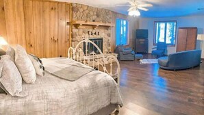 A bedroom with a bed, a stone fireplace, a ceiling fan, and a view of the outdoors.