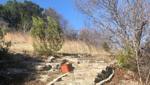 Una escalera de piedra que asciende con un contenedor rojo en la parte superior, rodeada de hierba seca y árboles.