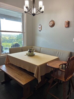 A dining area with a table set for two, a bench, a wooden chair, a chandelier, and a window with a view of greenery.