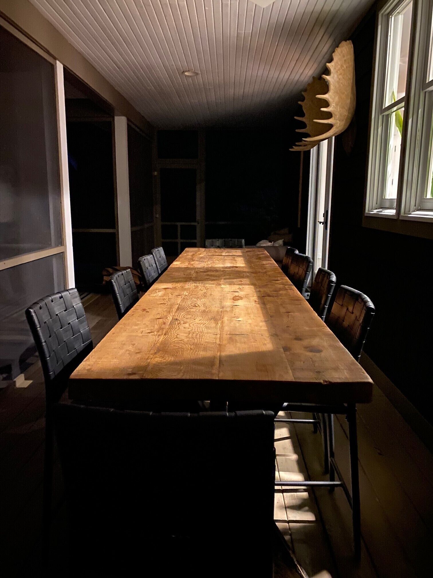 Une longue table en bois, entourée de chaises noires, se trouve dans une pièce à la lumière tamisée, dotée d’un plafond en pente et d’une grande fenêtre sur le côté droit.