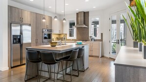 Una cocina moderna con una isla central, taburetes de bar, electrodomésticos de acero inoxidable y un ventanal amplio.