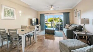 Un grand espace de vie comprenant une table à manger, des chaises, un canapé et un téléviseur. Un balcon offre une vue sur la verdure.