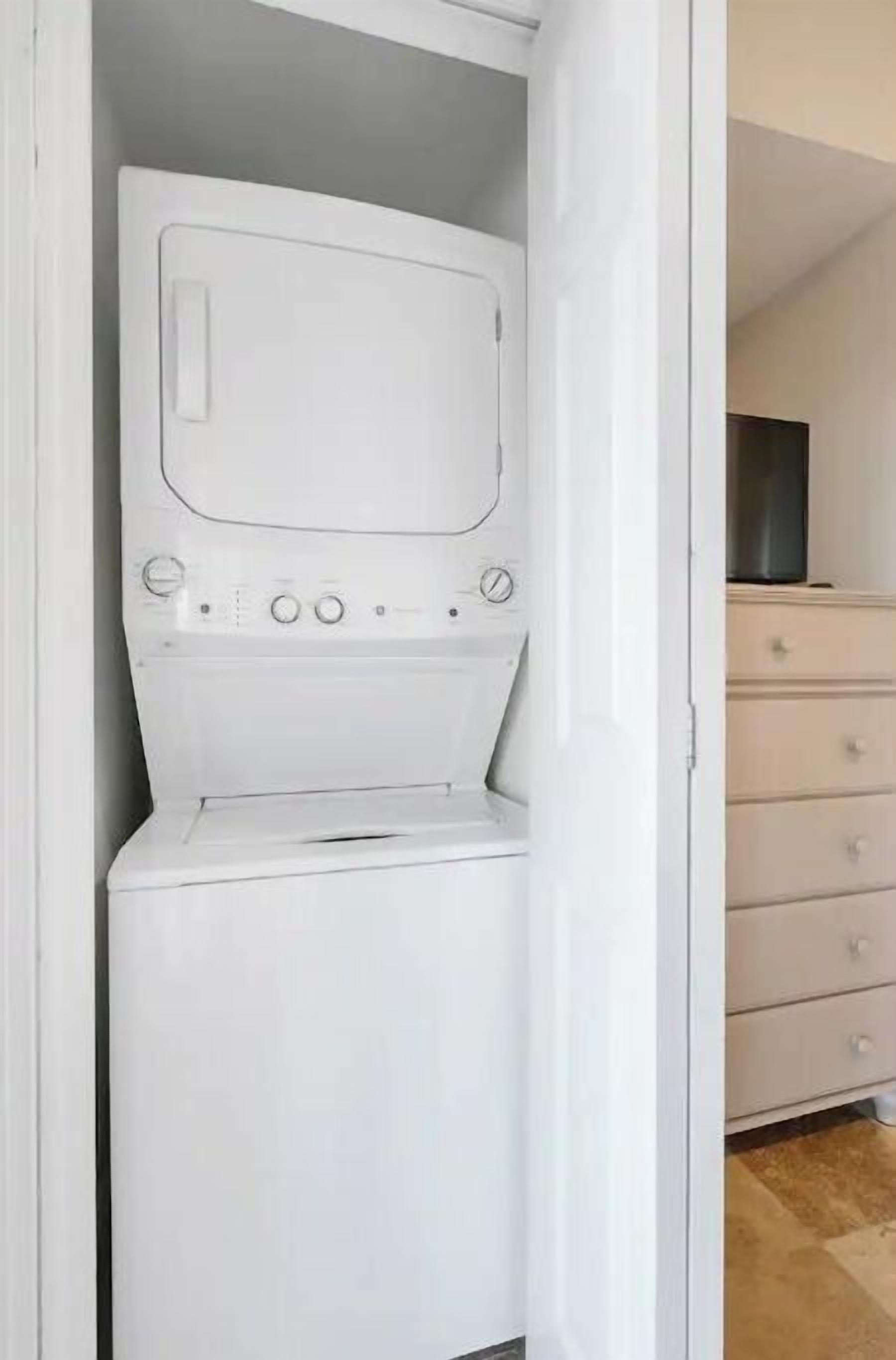 Máquina de lavar branca num armário, com uma televisão sobre uma cômoda ao fundo.