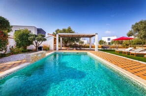 Une piscine d’un bleu éclatant, agrémentée d’une pergola et de chaises longues, entourée de verdure et de bâtiments modernes.