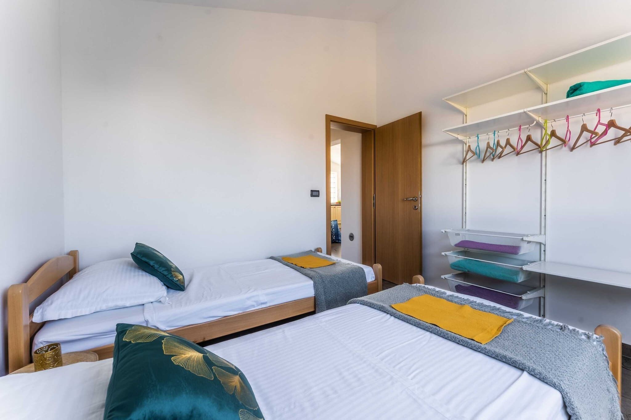 A room with two single beds, a wooden wardrobe, and a white shelf with colorful hangers.