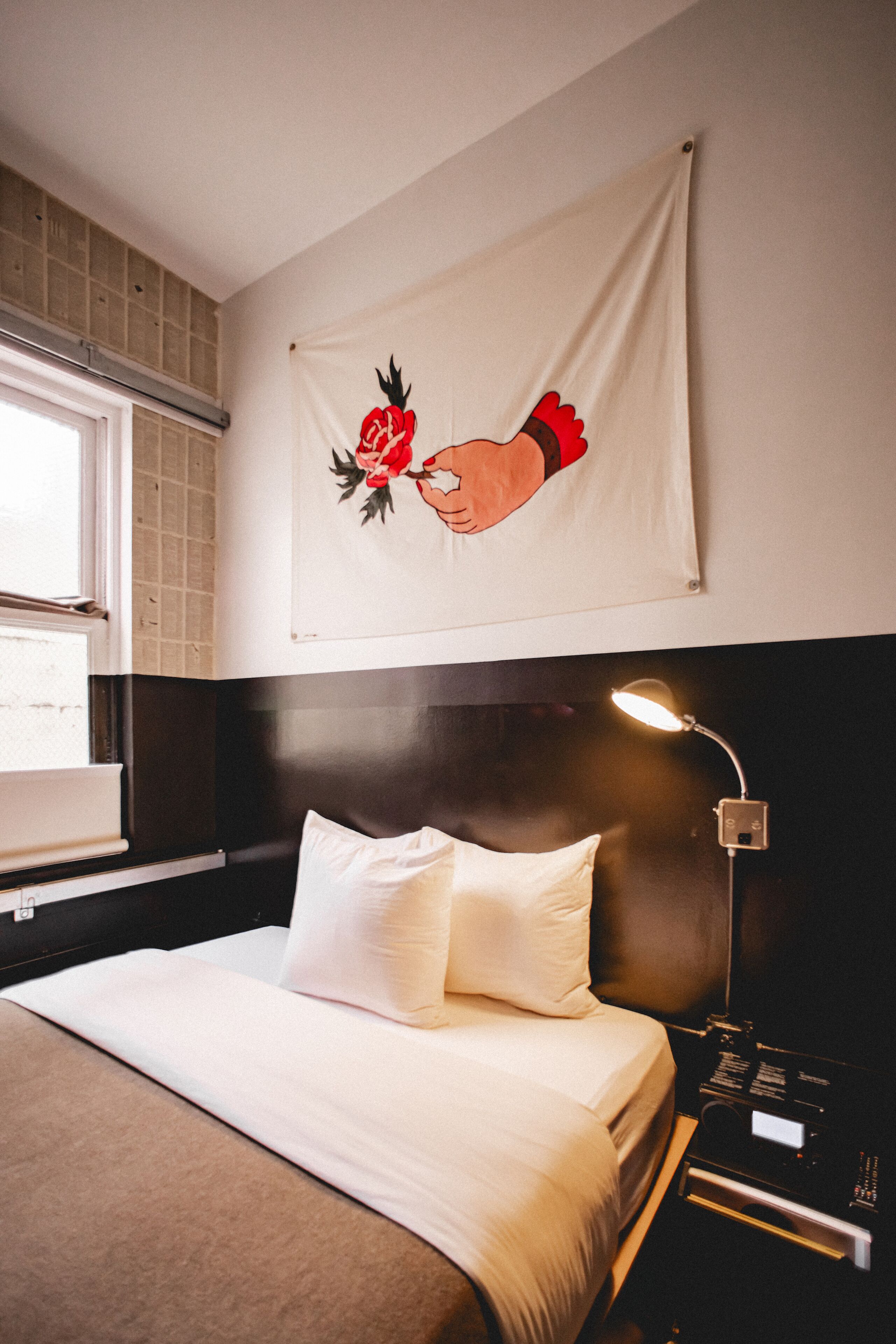 A hotel room with a bed, a nightstand with a lamp, a window, and a wall tapestry.