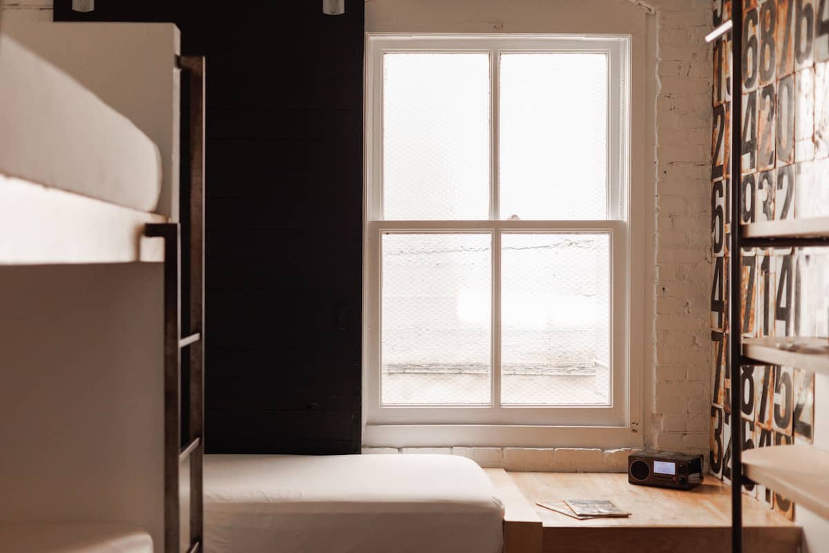 Een kleine, zwak verlichte kamer met stapelbedden, één raam en een boekenplank vol boeken.