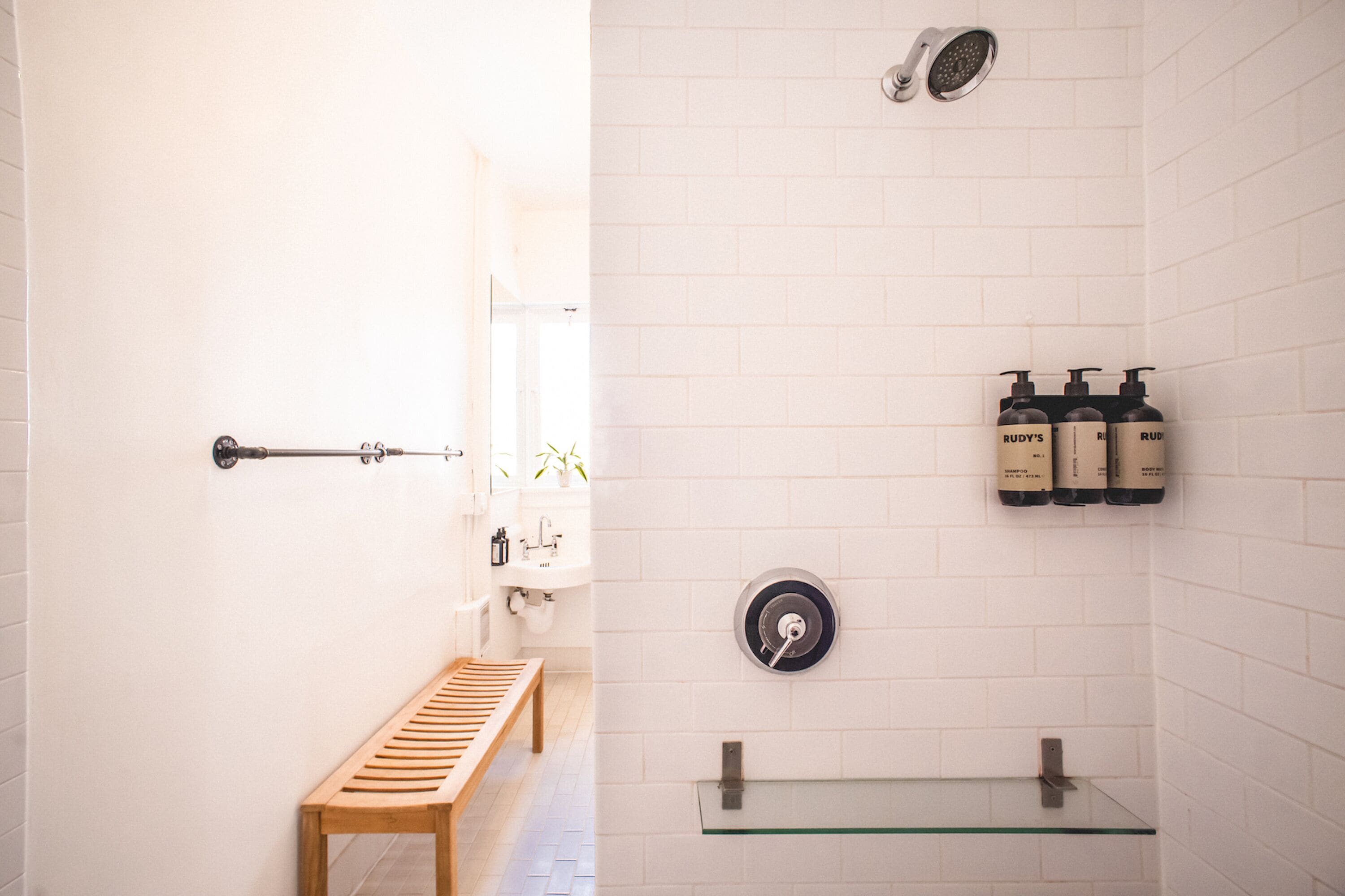 A bathroom with a wooden bench, a glass shelf, a toilet, and three bottles of shampoo on the wall.