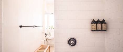 A bathroom with a wooden bench, a glass shelf, a toilet, and three bottles of shampoo on the wall.