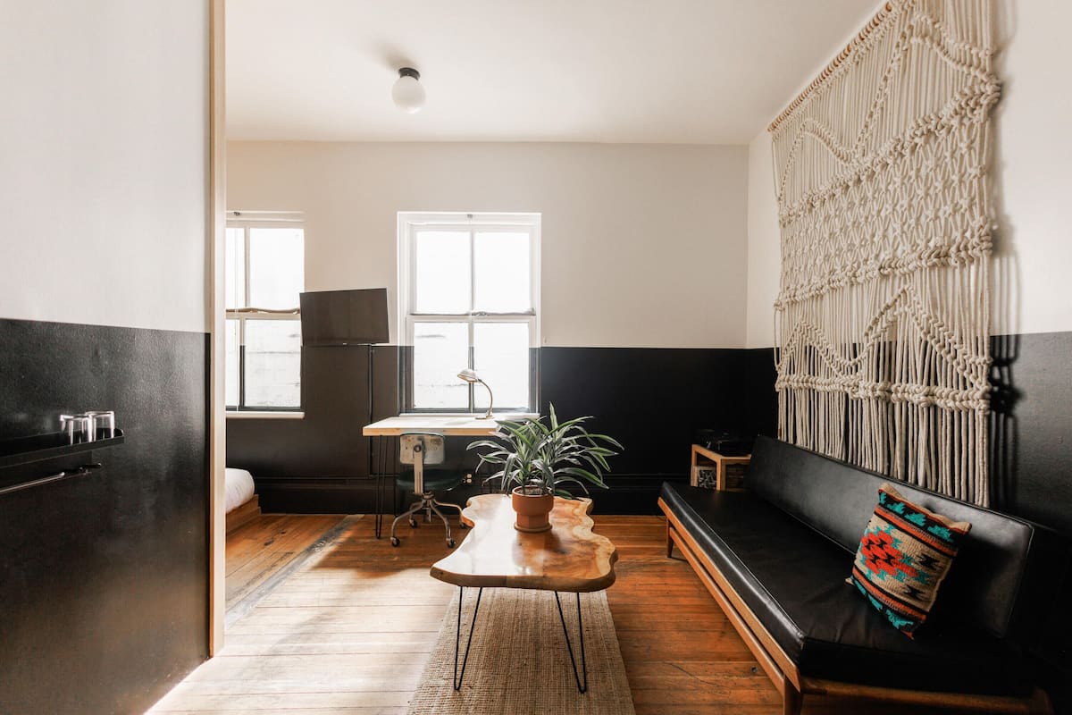 Een moderne woonkamer met een houten salontafel, een zwarte bank met een kleurrijk kussen, een macramé wanddecoratie en een potplant.
